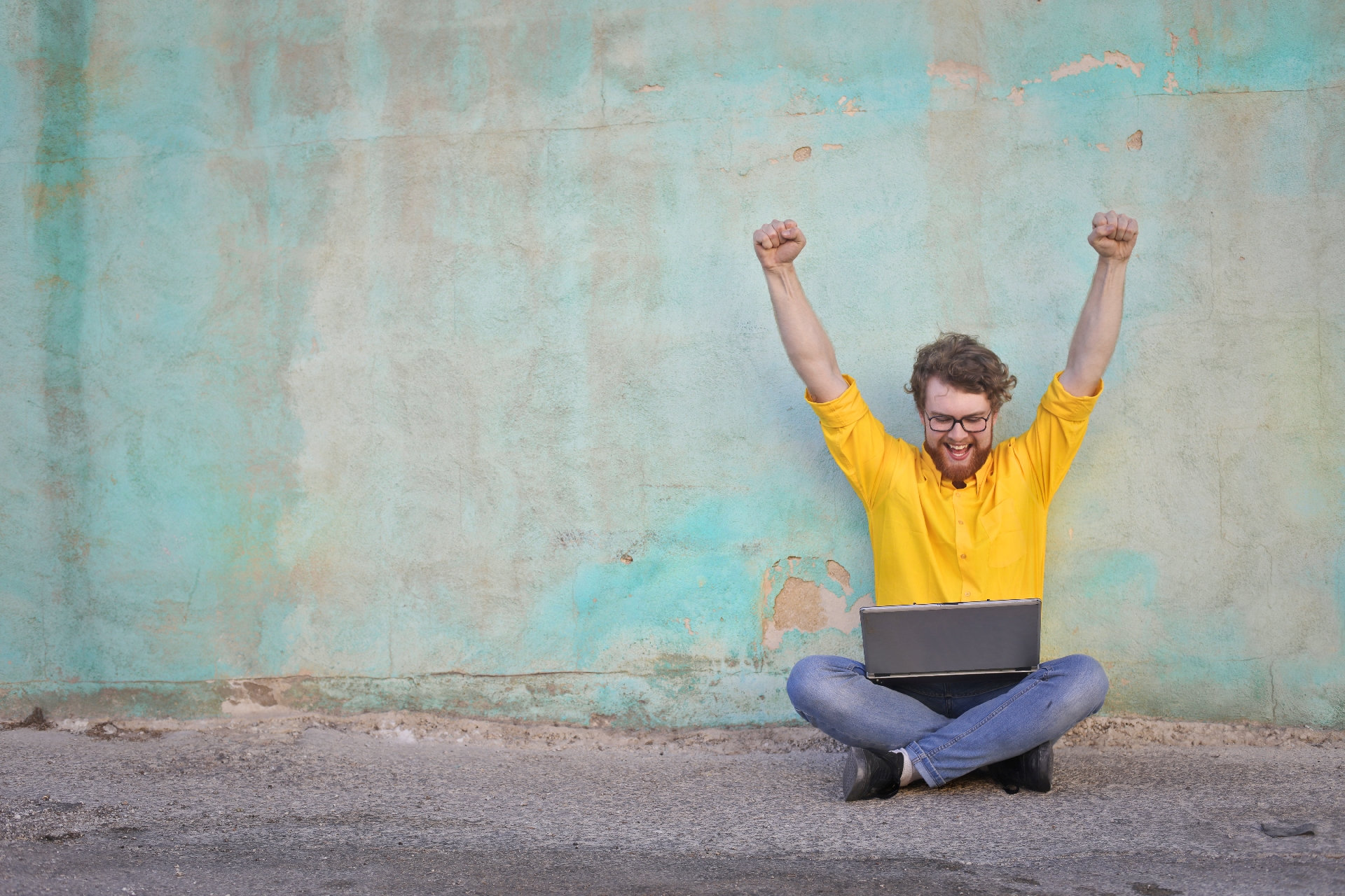 Bärtiger junger Mann mit Brille in sonnengelbem Hemd und Jeans sitz im Schneidersitz auf der Straße und reißt nach einem Blick auf seinen Laptop jubelnd die Arme hoch, Foto: AdobeStock/olly