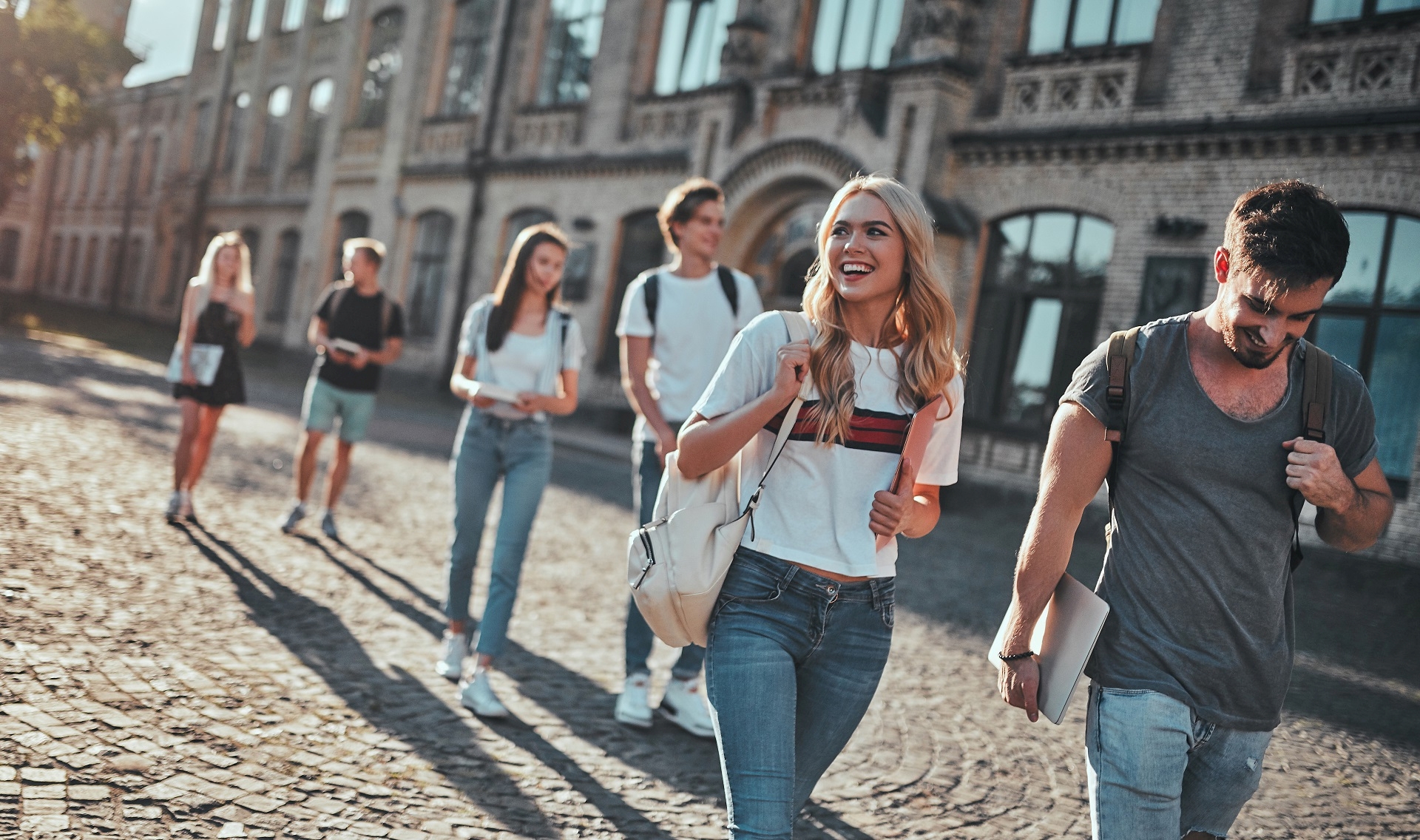 Fröhliche Gruppe junger Studierender mit Rucksäcken und Büchern unter dem Arm vor ihrer Universität, Foto: AdobeStock/Vasyl